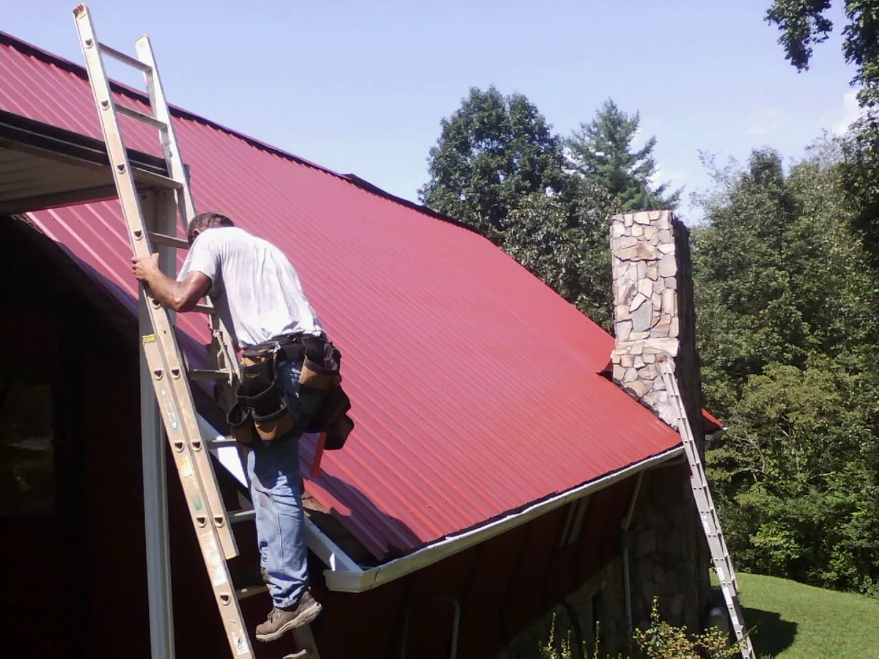 Roofer on a ladder beside a red metal roof and stone chimney — Soffit & Fascia Repair by Roofing Brothers in Toulon